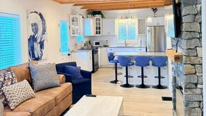 Un intérieur moderne avec un plafond en bois, un mur d’accent en pierre, une cuisine avec des meubles blancs et un coin salon comprenant un canapé et des tabourets de bar.