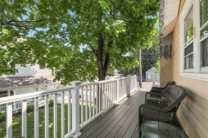 Una terraza de madera con barandas blancas, muebles de mimbre y un árbol que da sombra.