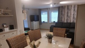 A dining area with a white table, wicker chairs, a vase with flowers, a television, and a sofa in the background. - Komfortable Ferienwohnung in Wenkendorf (Fehmarn)