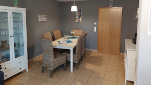 A dining area with a table, wicker chairs, a cabinet with glass doors, a wooden door, and a wall clock. - Komfortable Ferienwohnung in Wenkendorf (Fehmarn)