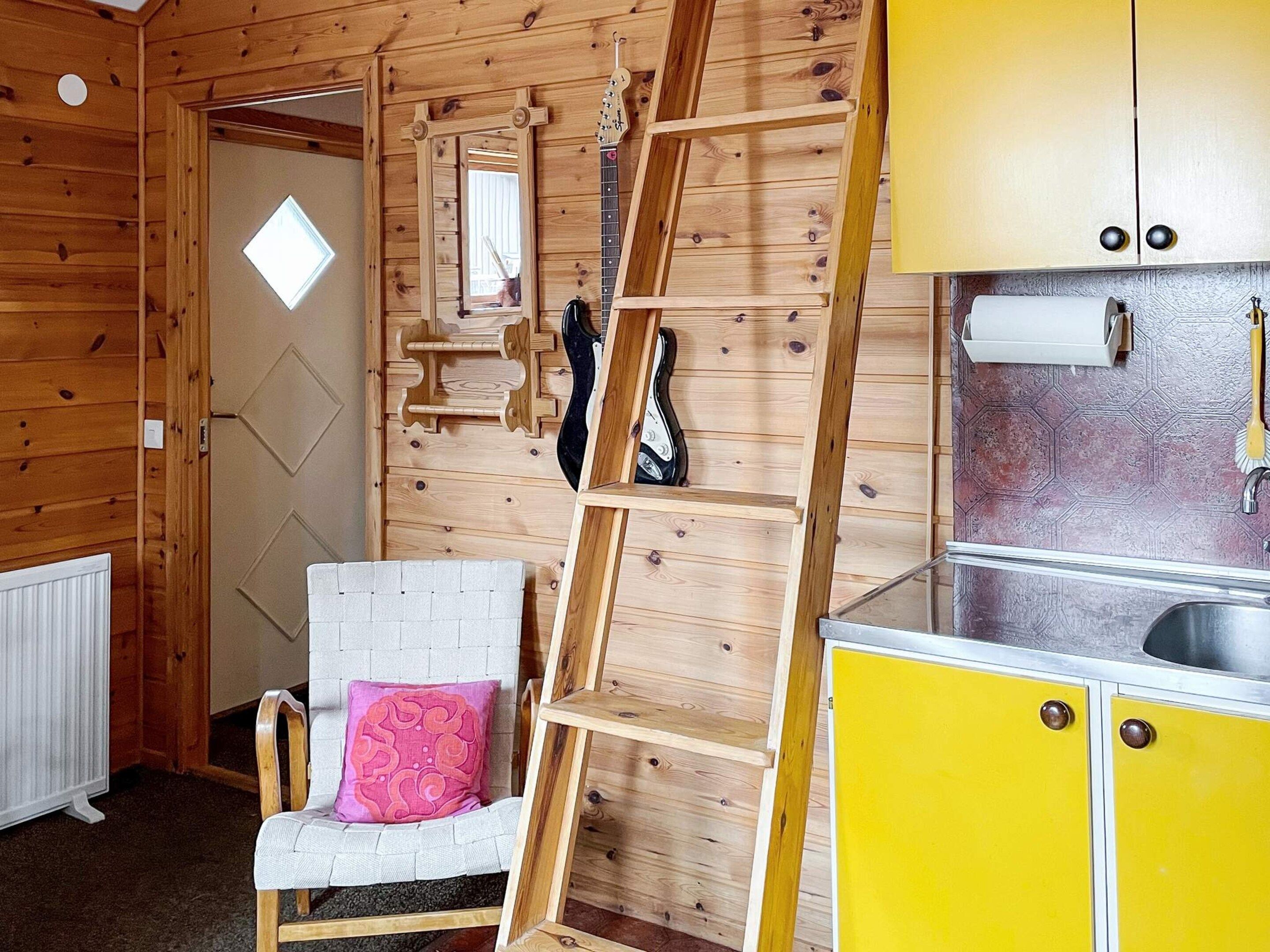 Interior de una cabaña de madera con una silla blanca, un cojín rosa, una escalera y armarios amarillos.
