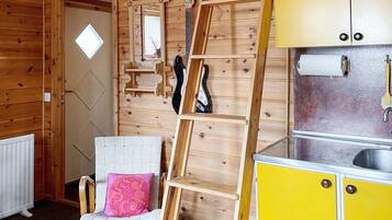 Interior de una cabaña de madera con una silla blanca, un cojín rosa, una escalera y armarios amarillos.