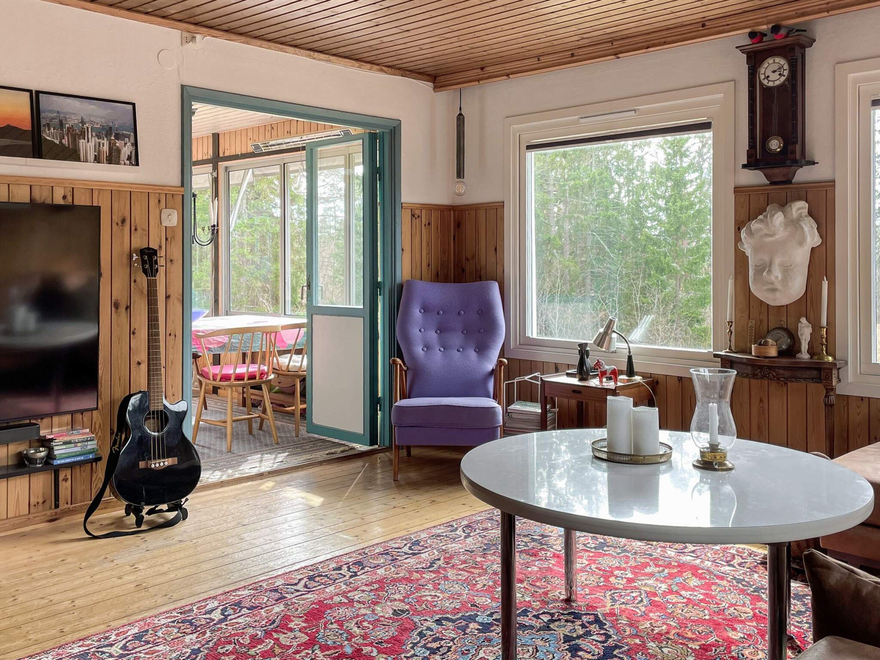 Una sala de estar acogedora con techo de madera, un ventanal grande, un sillón morado, una mesa redonda y un televisor.