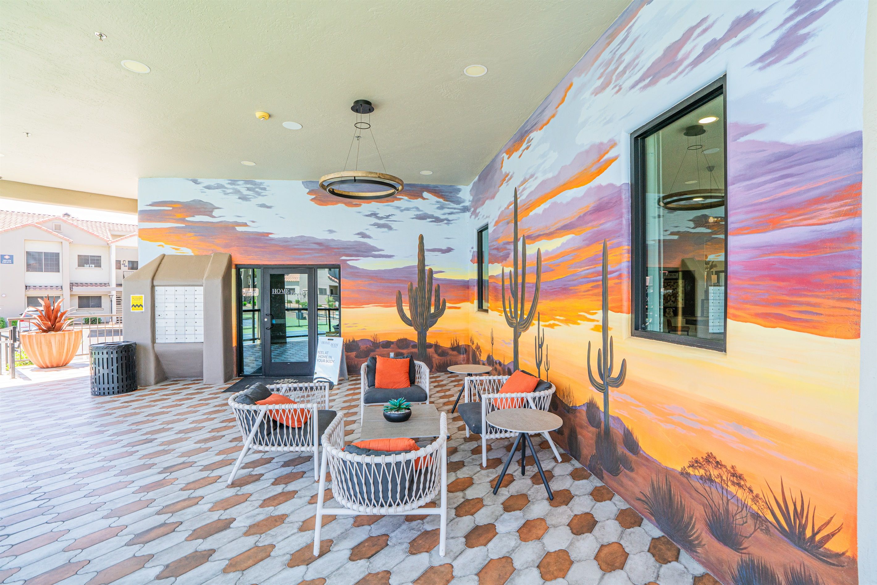 An outdoor seating area with wicker chairs and a table, a mural of a desert landscape, and a building entrance.