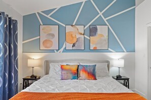 A hotel room with a bed, two bedside tables, a nightstand, a blue curtain, and abstract wall art.