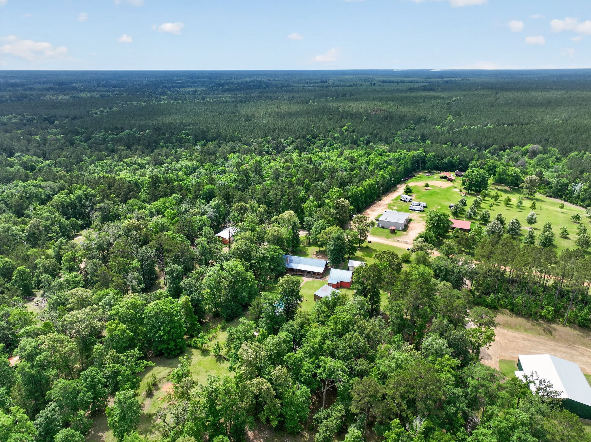 An aerial view of a rural property with scattered buildings, a large open grassy area, and dense forest surrounding it.