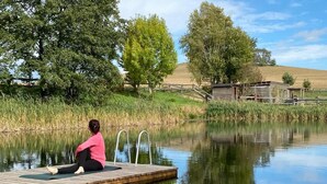 A person sitting on a dock by a calm body of water, surrounded by trees and a wooden structure in the background. - Duplex Flat With a Fireplace in a Historic Vaulted Building (Grossharthau)