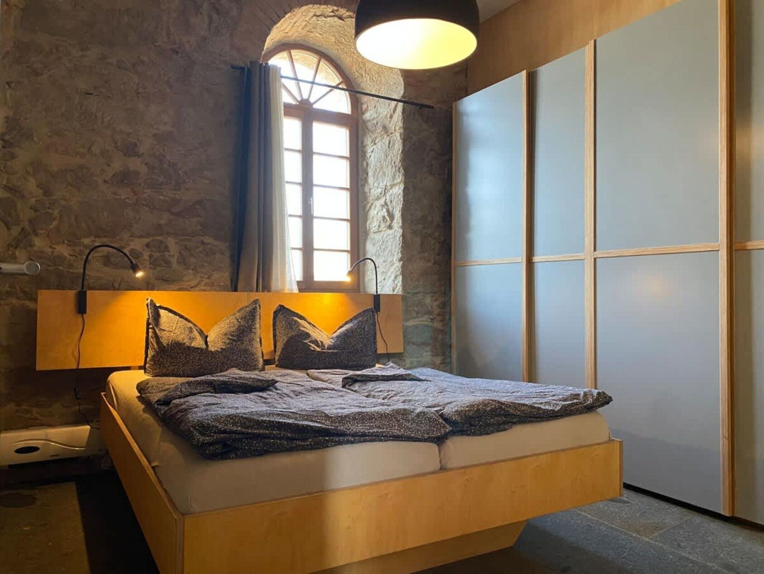 Un dormitorio moderno con pared de piedra, estructura de cama de madera, ventana grande y puerta corredera.