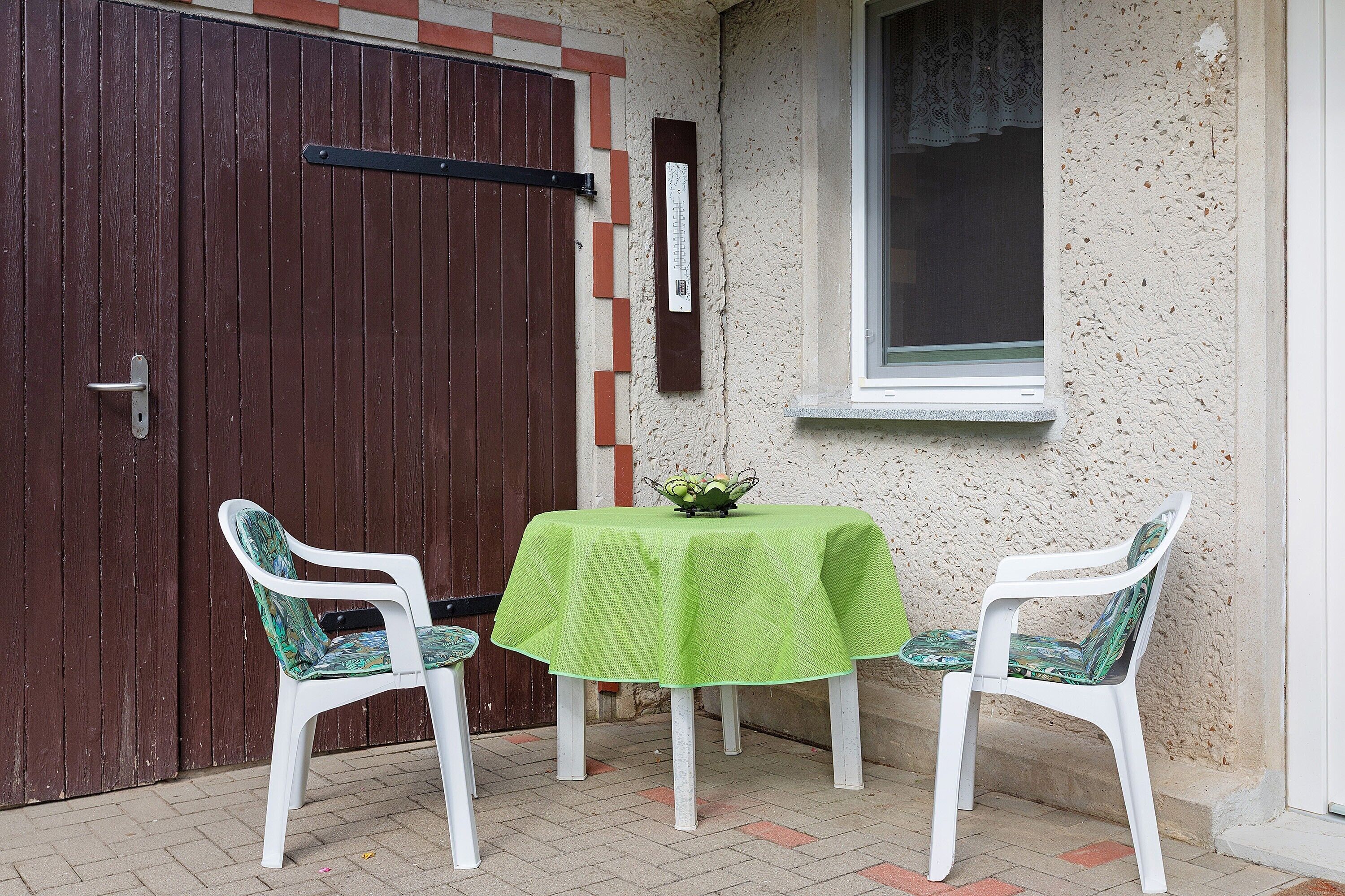 Ein kleiner Sitzbereich im Freien mit einem Tisch und zwei Stühlen, einer Tür und einem Fenster.