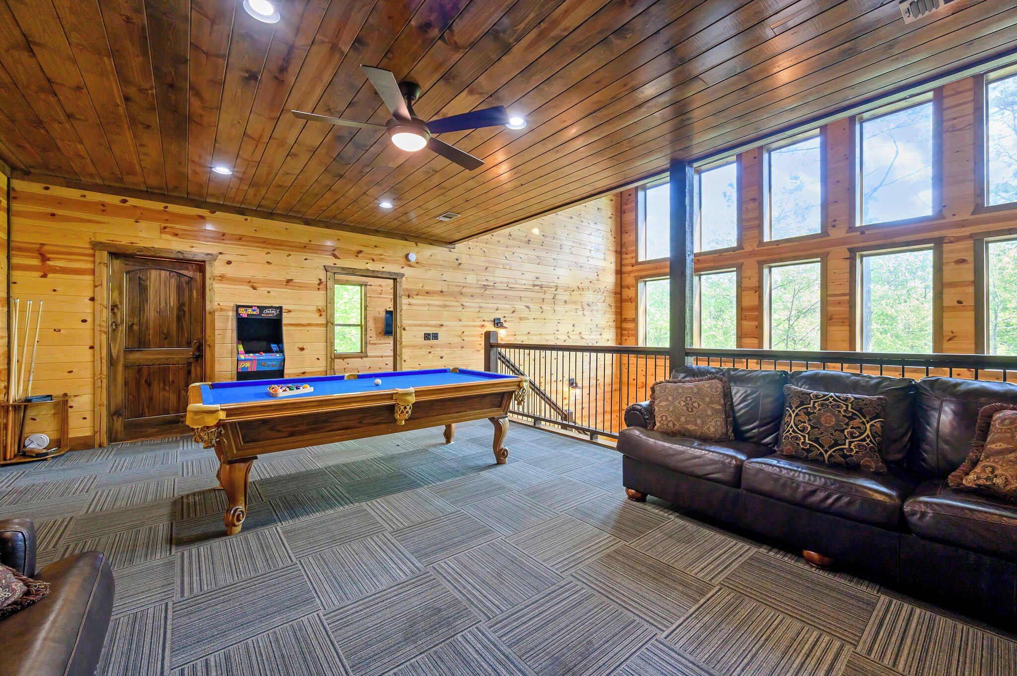 A wooden cabin interior with a pool table, leather sofa, and large windows.
