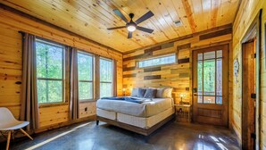 Une chambre dans une cabane en bois, avec un lit, un ventilateur de plafond et de grandes fenêtres.