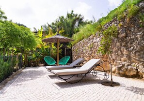 A paved outdoor area with lounge chairs and a small table, surrounded by greenery and a stone wall. - Carob Tree House - Paderne, Albufeira (Albufeira)