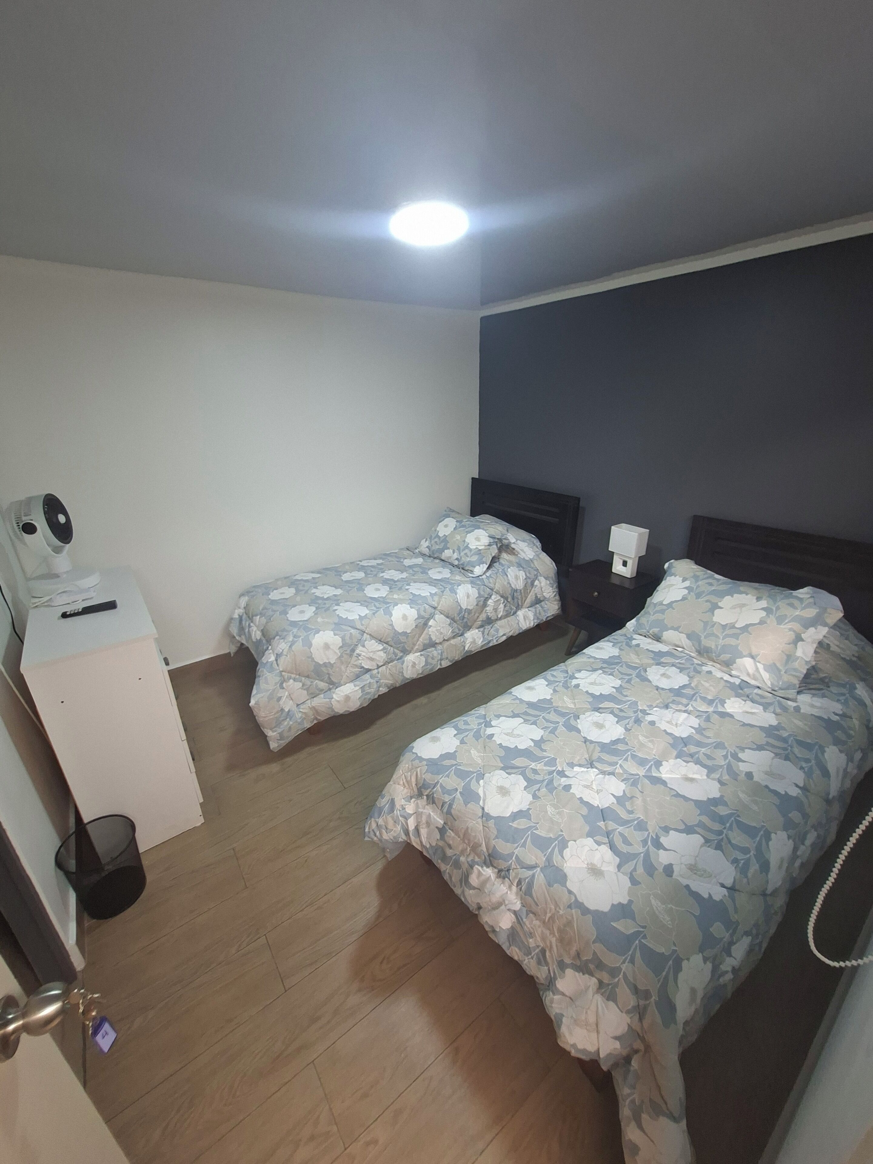 A room with two beds, a desk, a chair, a trash bin, and a wall-mounted light fixture.