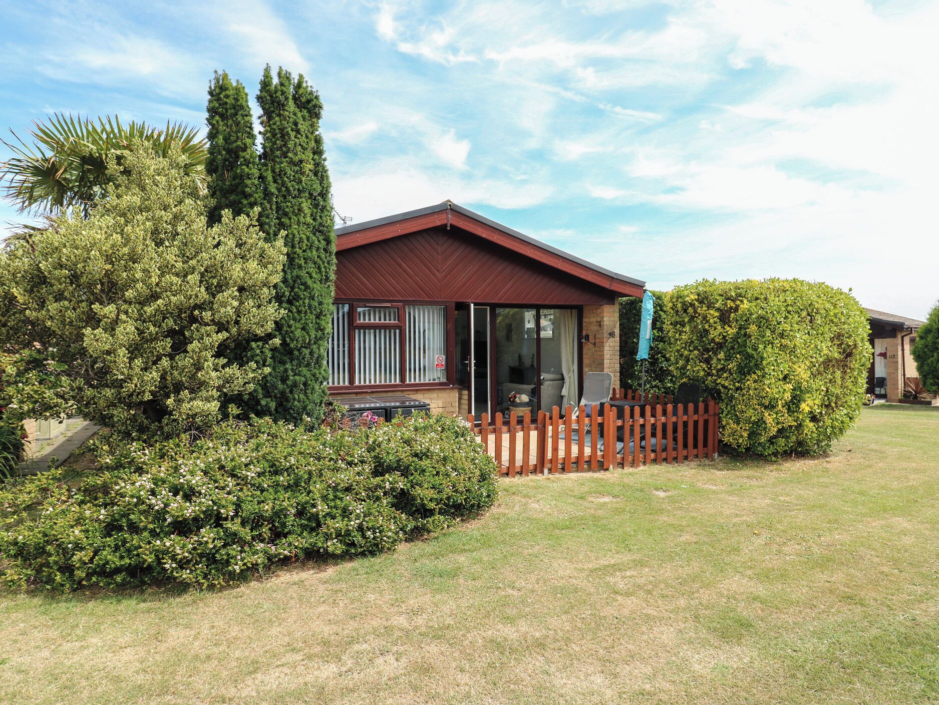 A single-story house with a red roof, a fenced yard, and a variety of greenery including a large bush and a tree.