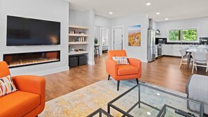 A modern living room with a fireplace, orange armchairs, a flat-screen TV, and a kitchen in the background. - Fox Den | 4 Suite Retreat + EV | 10 Mins to AVL (Asheville)