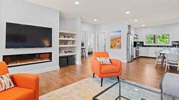 A modern living room with a fireplace, orange armchairs, a flat-screen TV, and a kitchen in the background.