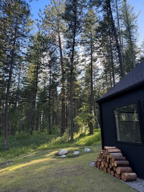 A cabin with a stack of firewood and a clear view of a forested area. - Your Mountain Modern Glacier Escape in the Pines (Columbia Falls)