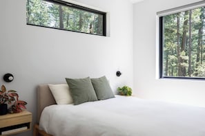 Un dormitorio con una cama, dos ventanas, una mesita de noche con una planta y una lámpara fijada a la pared.