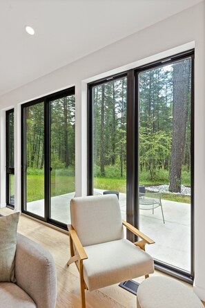 A modern interior with large windows, a sofa, and a chair facing a forest view. - Your Mountain Modern Glacier Escape in the Pines (Columbia Falls)