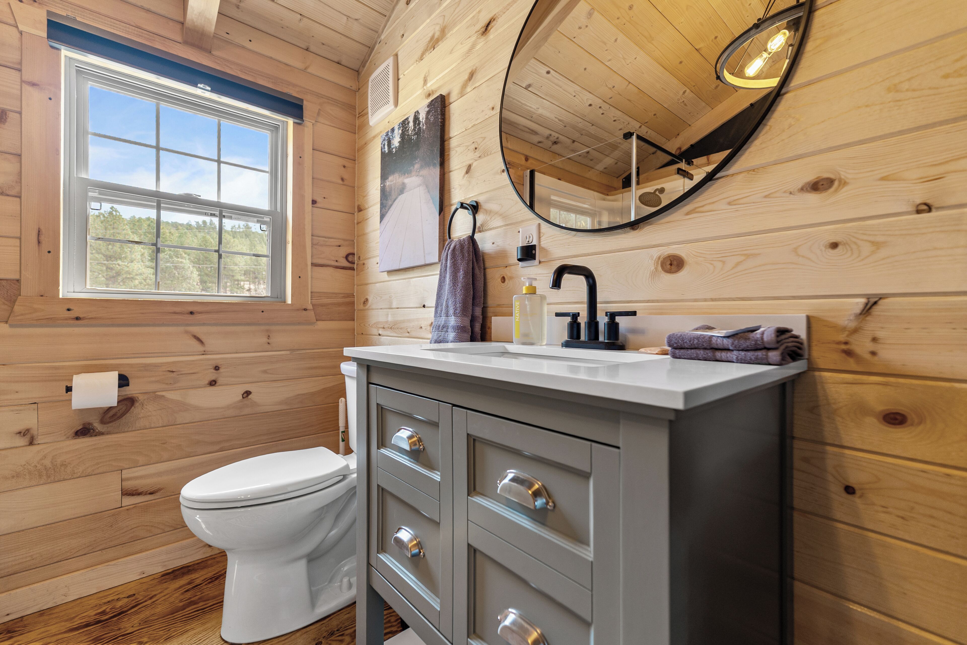 A wooden cabin bathroom with a toilet, vanity, mirror, and window.