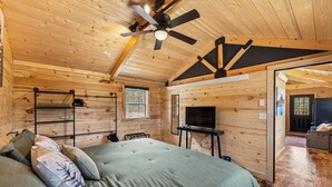 A wooden cabin interior with a bed, a ceiling fan, a television, and a fireplace.
