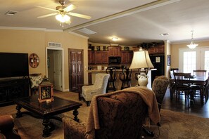 A living room with a television, a coffee table, and a fireplace. There is a kitchen area with wooden cabinets and a dining table.