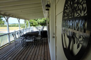 A covered outdoor seating area with tables and chairs, a white wall with a metal sign, and a view of a green landscape.