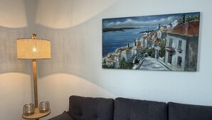 A living room with a dark-colored sofa, a lamp with a cylindrical shade, and a painting of a coastal town.