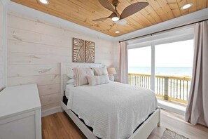 Ein Schlafzimmer mit einem Bett, einem Nachttisch, einem Deckenventilator und einem großen Fenster mit Blick auf den Strand.