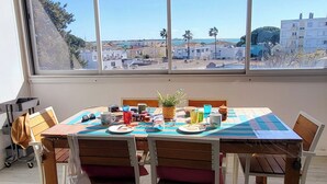A dining area with a table set for two, a view of the city through large windows, and a bag on the floor. - Le plein Sud entre Plage 🏖️ et Commerces (Le Grau-du-Roi)