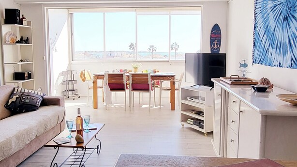A modern living room with a sofa, coffee table, and TV. There's a dining area with chairs and a table, and a large window offering a view. - Le plein Sud entre Plage 🏖️ et Commerces (Le Grau-du-Roi)