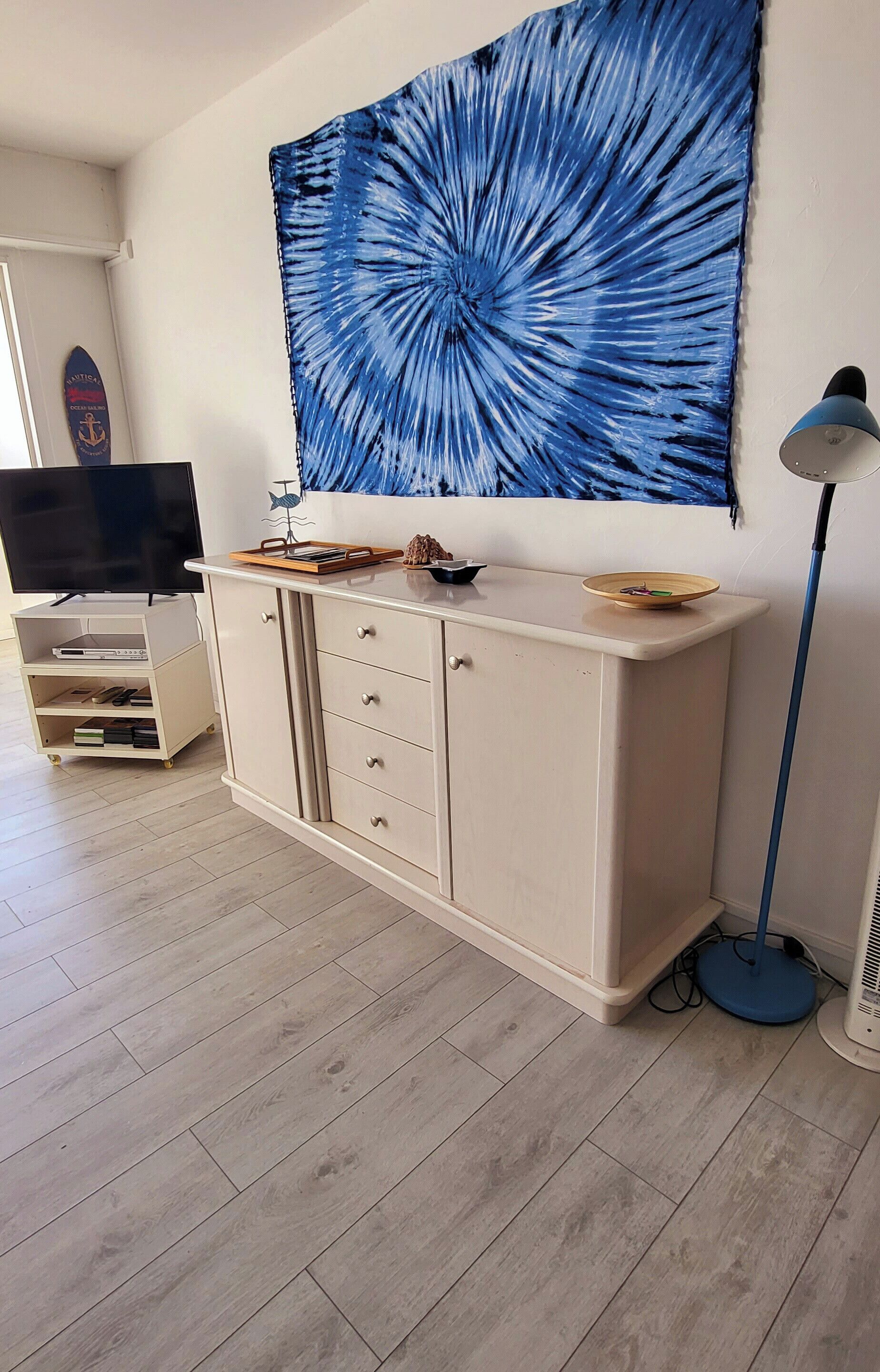 Ein Zimmer mit Fernseher auf einem Schrank, einer Kommode mit Schubladen, einem großen blauen Kunstwerk an der Wand und einer Stehlampe.