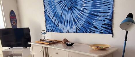 Ein Zimmer mit Fernseher auf einem Schrank, einer Kommode mit Schubladen, einem großen blauen Kunstwerk an der Wand und einer Stehlampe.
