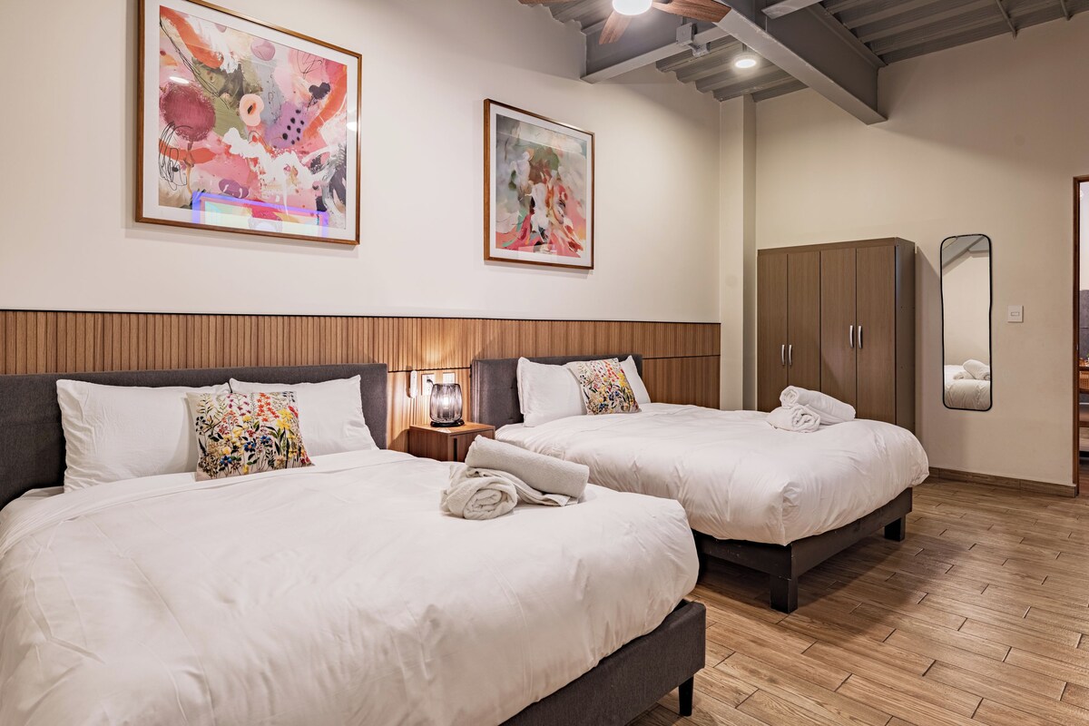 Habitación de hotel con dos camas, cabecero de madera, obras de arte en las paredes, un armario y un espejo.