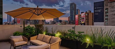 A rooftop terrace with wicker furniture, an umbrella, and potted plants against a city skyline.