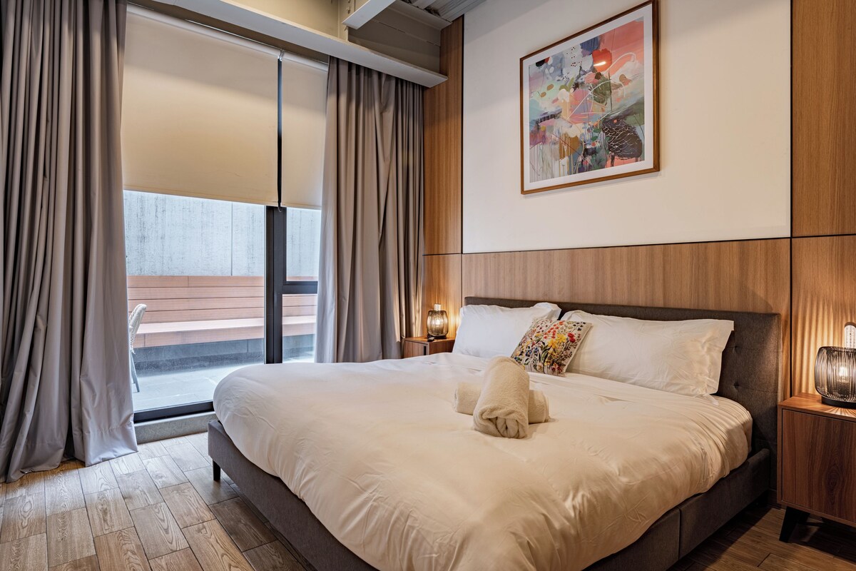 Habitación de hotel con una cama grande, una ventana con cortinas, una mesita de noche con lámpara y un cuadro en la pared.