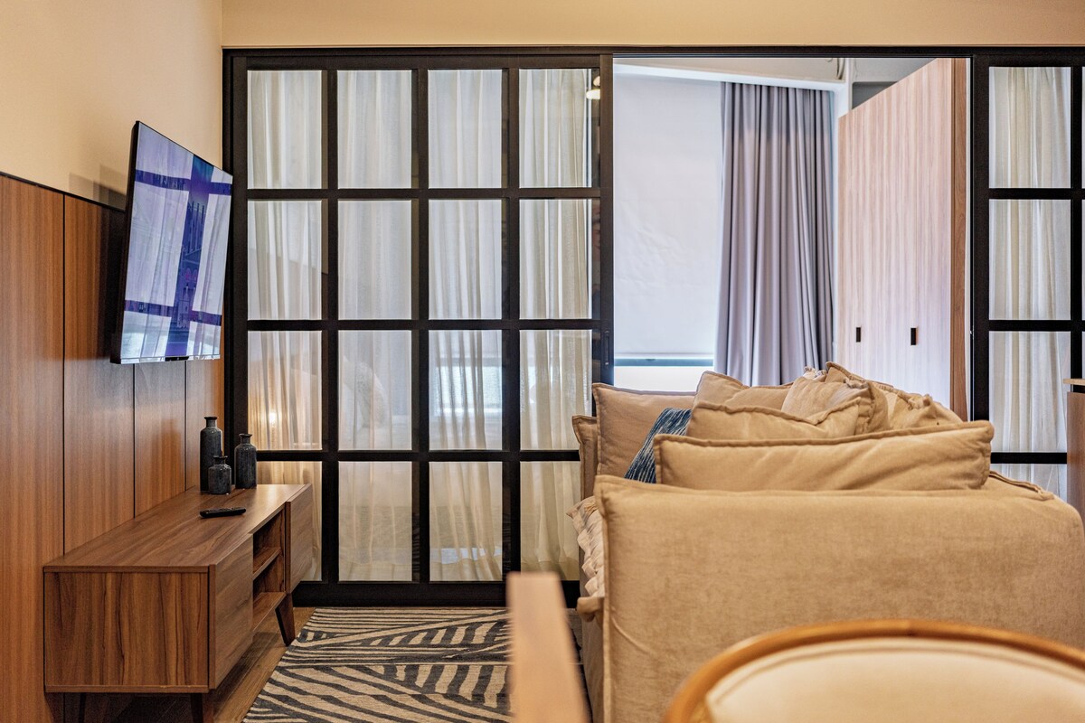 Habitación de hotel con una cama, un televisor, un tocador de madera y vistas a través de un ventanal con cortinas.