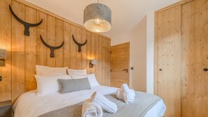 Quarto com cabeceira de madeira, cama com roupa de cama branca, manta cinza e toalhas dobradas sobre a cama.