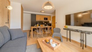 Uma sala de estar moderna com sofá, mesa de jantar com cadeiras, área de cozinha com armários e eletrodomésticos, e uma TV fixada na parede.