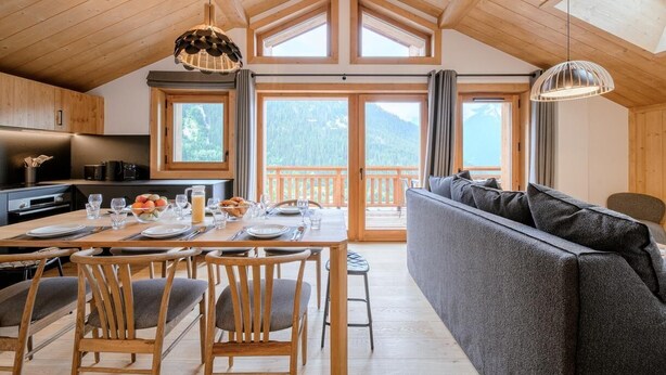 Una habitació amb sostre alçat, taula de dinar per a quatre places, sofà i vista a un paisatge montanyós a través de grans finestres.
