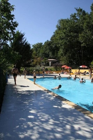 Ein Schwimmbeckenbereich mit Schwimmern und Spaziergängern, umgeben von Bäumen und Sonnenschirmen.