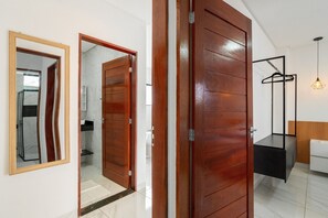 Une piĂšce avec une porte en bois, un miroir, un luminaire suspendu et un lit blanc.
