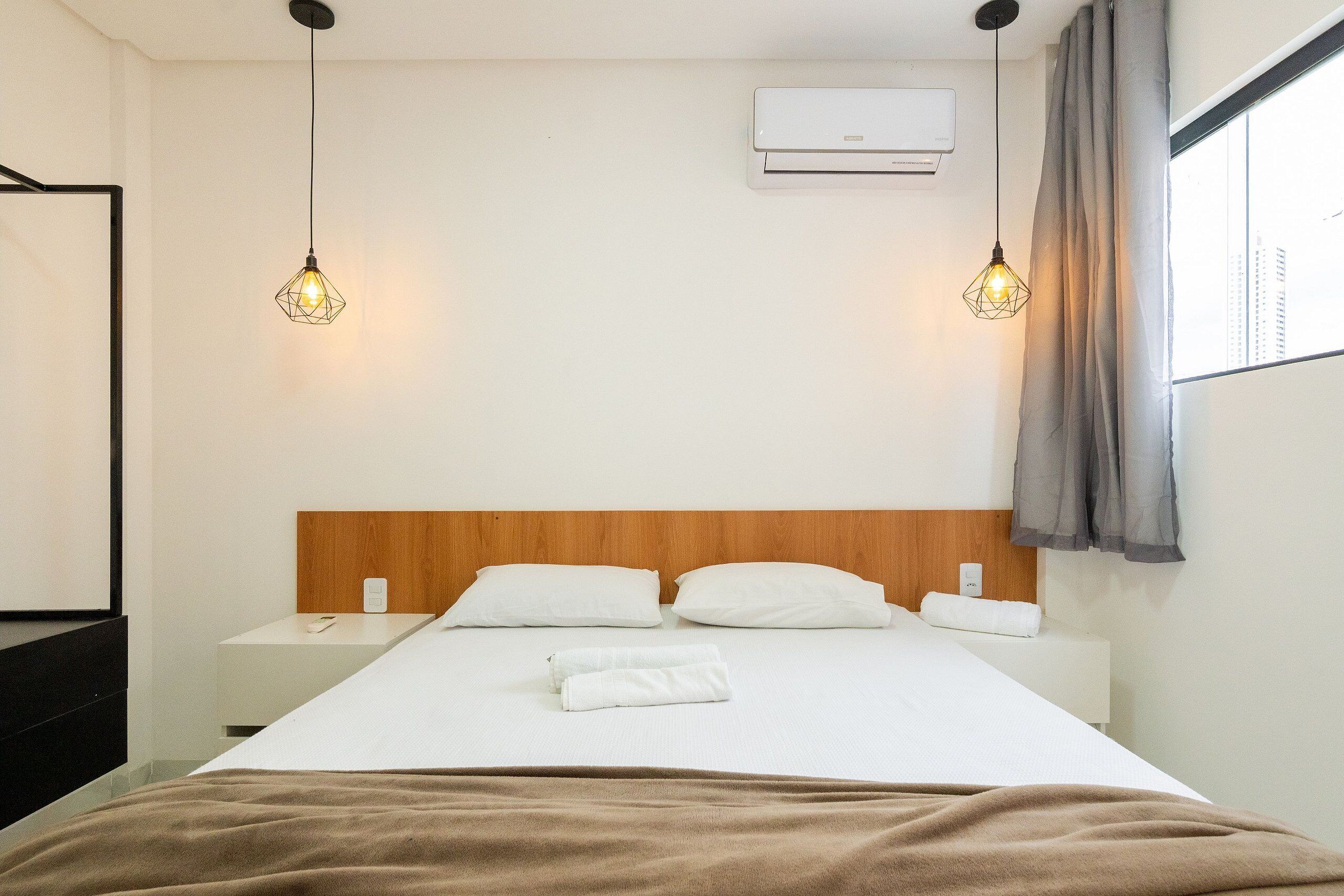 A hotel room with a bed, two bedside tables, a wooden headboard, an air conditioner, and two pendant lights.