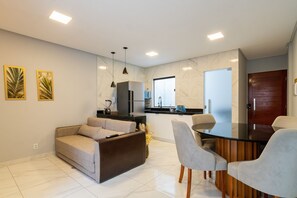 A modern kitchen with a refrigerator, microwave, and sink. There is a dining area with a glass table and chairs. A sofa is placed in the living area.