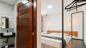Una habitación de hotel con puerta de madera, un baño con lavamanos y espejo, una cama con cabecero y una lámpara colgante.