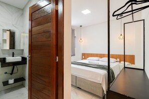 Quarto de hotel com porta de madeira, banheiro com pia e espelho, cama com cabeceira e luminária pendente.