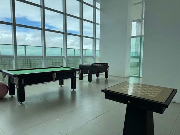 A pool table with a green felt surface, a foosball table, and a chess table in a room with large windows.