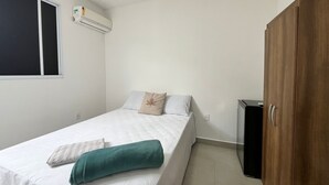 A single bed with white bedding and a teal towel, a wooden wardrobe, a small black cabinet, and a window with blinds. - Caminho das Bromélias Lodging – Caruaru (Caruaru)