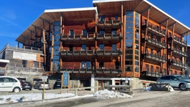 Un immeuble à plusieurs étages, doté d’une façade en bois et de balcons, situé dans une région enneigée, avec des voitures garées devant.