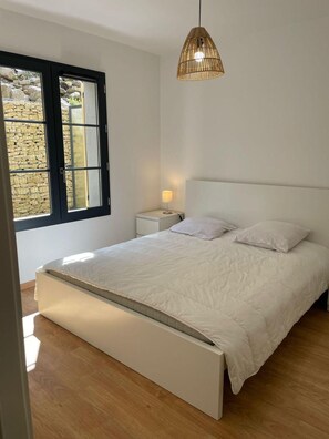 Ein Schlafzimmer mit Bett, Nachttisch, Lampe und einem Fenster mit Blick auf eine Steinmauer.